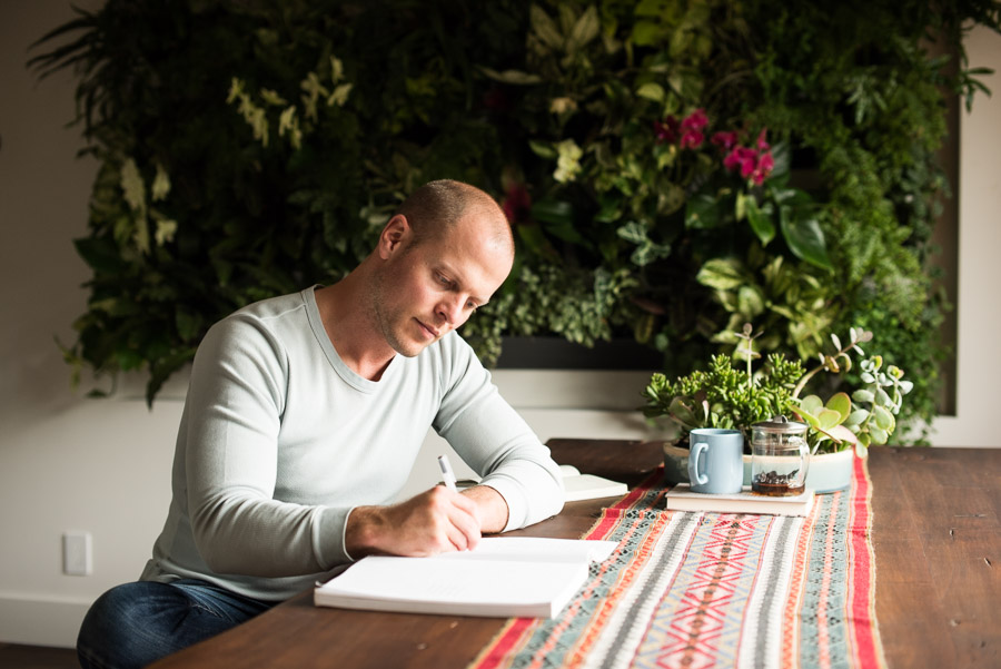 Tim Ferriss sitting at a table with plants behind him and writing in a notebook.