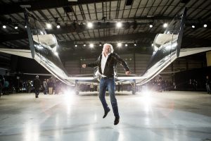 Richard Branson in an airplane hangar, jumping in the air in front of a futuristic looking airplane.