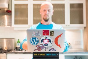 Tim Ferriss in his kitchen on his laptop that's perched on his books and decorated with San Francisco, WordPress, Thunderbirds, and Adopt stickers.