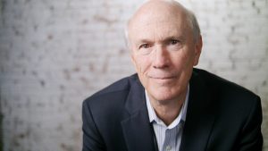 Frank Blake in front of an interior brick wall that has been painted white.