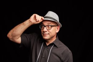 Cal Fussman in a checkered trilby, standing in front of a black backdrop.
