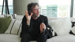 Neil Gaiman sitting on a sectional couch and holding a microphone.
