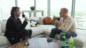 Tim Ferriss interviewing Neil Gaiman on a sectional couch before a large window overlooking Austin.