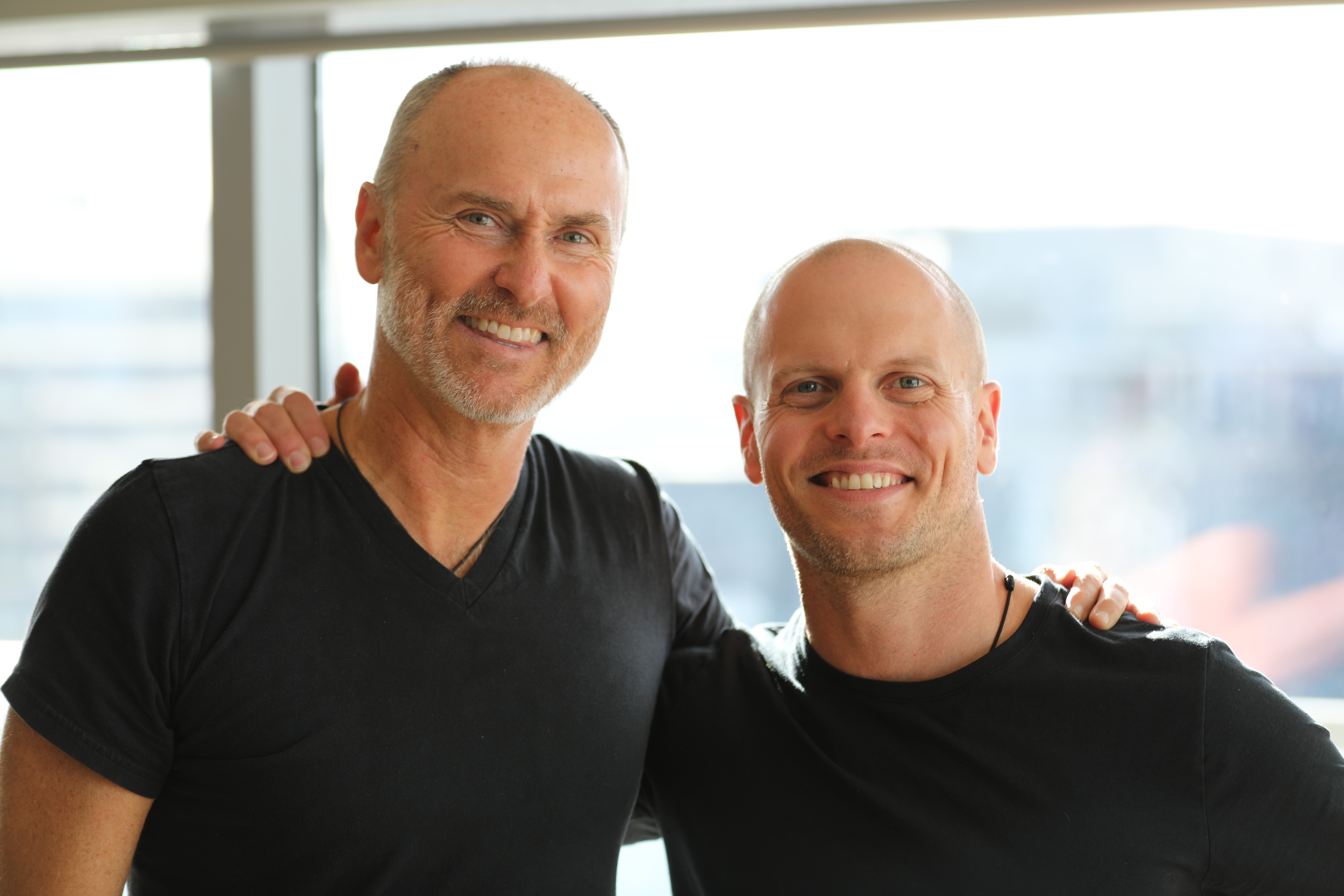 Tim Ferriss and Chip Conley standing in front of a window, smiling.