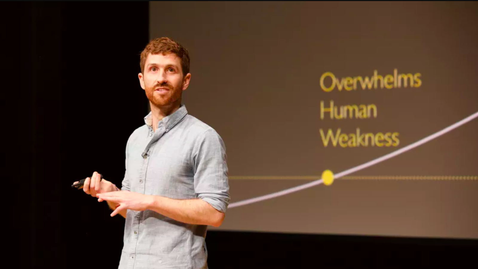 Tristan Harris onstage giving a presentation with a projected slide behind him that says, "Overwhelms Human Weakness."