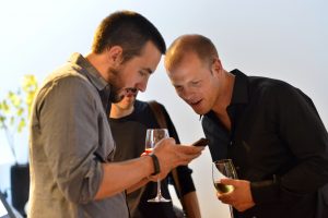 Tim Ferriss and Kevin Rose holding glasses of wine and looking at Kevin's smartphone.