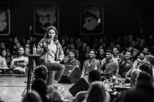 Whitney Cummings on a circle in the round stage doing stand-up for an all male audience.