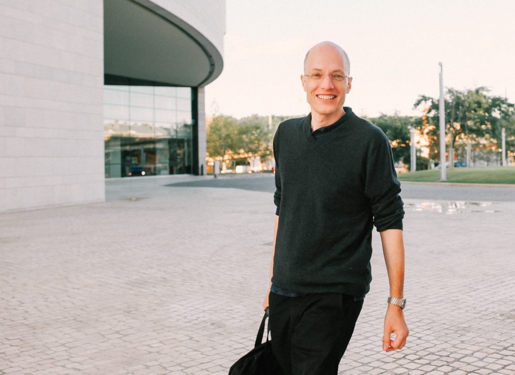 Alain de Botton carrying a tote bag in front of a modern, concrete building.