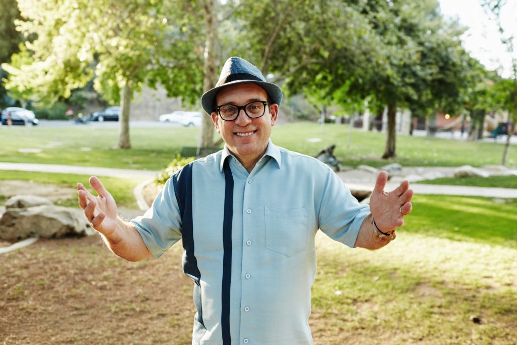 Cal Fussman, smiling and standing in a park with his arms open.