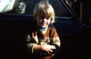 Tim Ferris at around 4 years old, arms wrapped around himself, standing in front of a car.