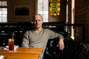 Tim Ferriss sitting on a black, leather, tufted couch in front of a wood table.