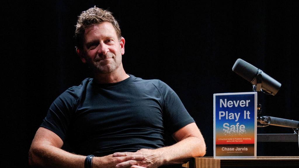 Chase Jarvis siting at a podcast desk with a microphone next to his new book Never Play It Safe.