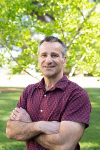 Dr. Keith Baar outside in a short sleeve maroon polo, in front of grass and deciduous trees on a sunny day.