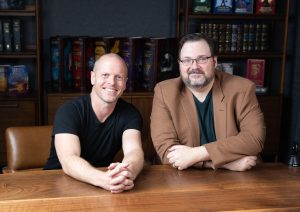 Tim Ferriss and Brandon Sanderson at Sanderson's headquarters, sitting at a large conference table surrounded by Sanderson's epic novels in special-edition illustrated bindings.