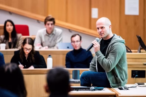 Tim Ferriss sitting on a desk in a small lecture hall at Harvard Business School and addressing students.