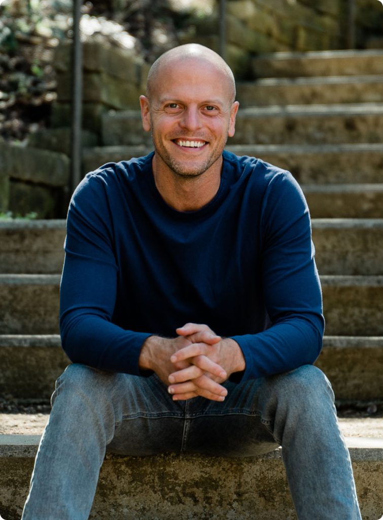 Tim Ferriss in a blue long-sleeve shirt and jeans sits on outdoor stone steps with his hands clasped, looking at the camera.
