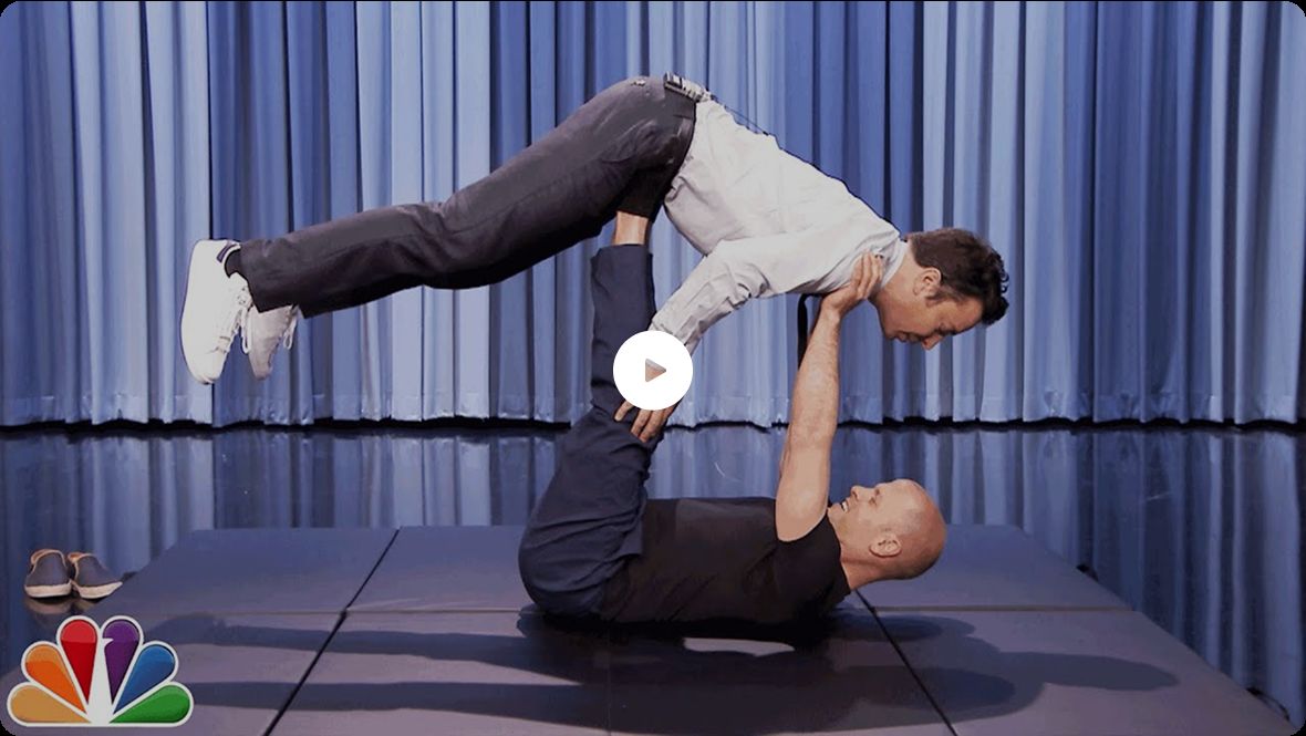 Tim Ferriss and Jimmy Fallon perform an acro yoga pose on a stage; one man lies on his back with his legs raised, supporting the other man horizontally in the air with his feet. An NBC logo appears in the corner.